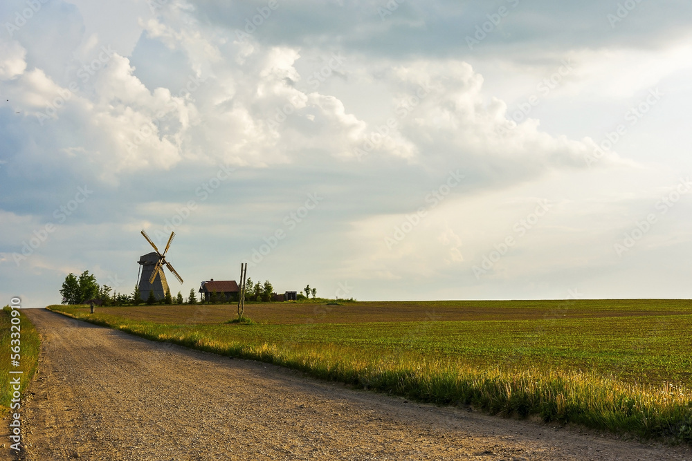 Fototapeta premium Dirt road to the lonely windmill and farmhouse located among the field during sunset