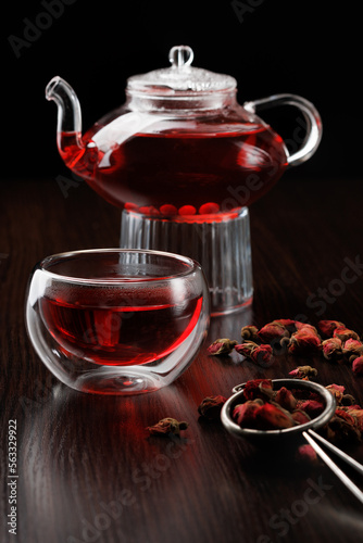 Glass teapot with red herbal tea on a black background. Side view selective focus. In the foreground is a glass thermos cup and dry tea leaves of hibiscus.