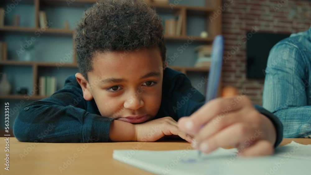 Tired exhausted sad upset fatigued little African American ethnic child boy kid son pupil schoolboy schoolchild lying on table desk writing homework boring class lesson drawing in notebook near father