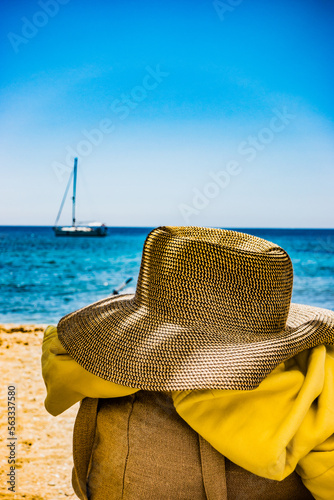 Un chapeau en paille est posé sur une chaise sur une plage  en été devant une mer bleu turquoise , on devine un voilier au loin
