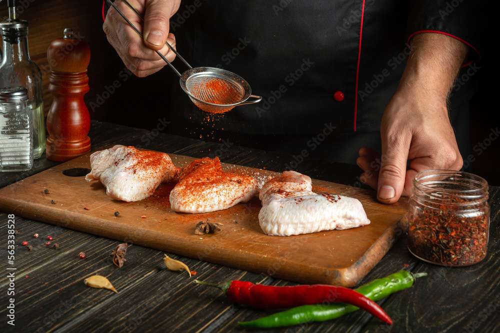 Professional cook adds paprika to chicken wings. Raw chicken wing in the kitchen of a restaurant or cafe on a cutting board with vegetables and spices