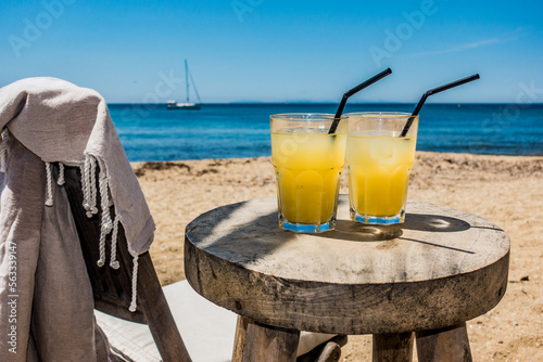 Des cocktails de fruits sont posés sur une jolie table en bois sur une plage magnifique en été, on devine un voilier mouillé au large .