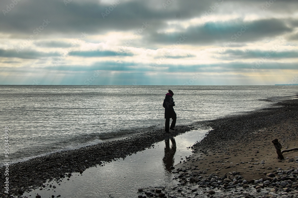 On a mostly cloudy Winter day in Wisconsin, a silhouetted individual walks along the Lake Michigan shoreline and is reflected in a small stream along the shore.