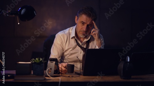 Businessman sitting at desk with laptop computer, working late in office, tired. Entrepreneur thinking, solving problem. Older, middle aged, mid adult, man in his 40s or 50s in shirt and tie.