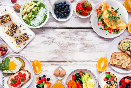 Healthy breakfast food frame. Overhead view on a white wood background. Table scene with omelette, nutritious bowl, toasts, granola bars, smoothie bowl, yogurts, fruit. Copy space.