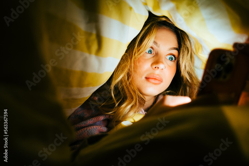 Fotografie Young amazed caucasian woman using smartphone, lies under a blanket on a bed at home in the evening