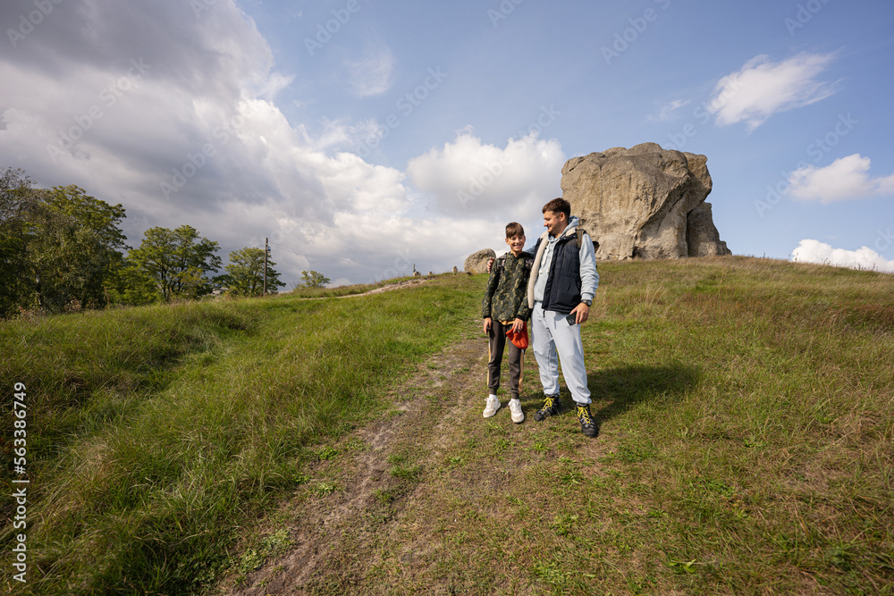 Son with father wear backpack hiking near big stone in hill. Pidkamin, Ukraine.