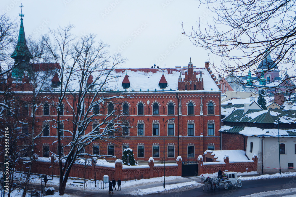 Obraz premium Krakow, Poland - December 20, 2022. Cityscape. Traditional European Old Town Street with Red Building (The Straszewski Palace) and a carriage with horses. European City Architecture.