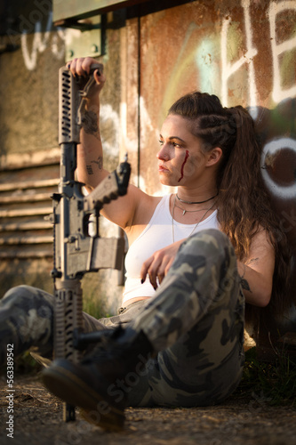 Caucasian young woman wearing camouflage battledress and holding a mockup rifle sits with her back against the front wall of bunker with graffiti.