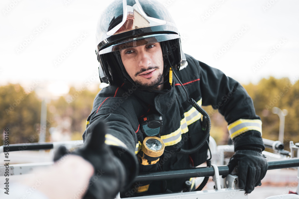 A young firefighter climbs on top of the basket of a fire truck cranes raising his arm to shake hands with a person in a building who is trapped.