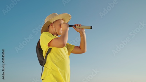 Child plays in nature in traveler. Boy in hat looks through spyglass in summer outdoors. Kid, son, dreams of discoverer of childhood fantasies. Childrens travel. Teenager watches through spyglass