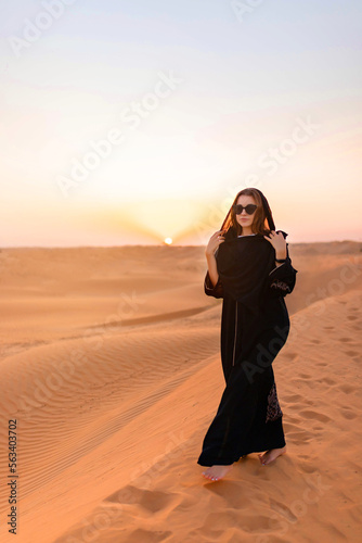 Beautiful mysterious woman in traditional arabic black long dress stands in the desert on sunset