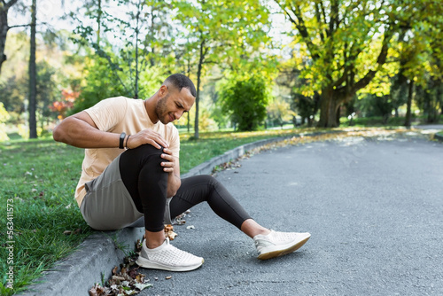 Man injured leg while jogging, african american man sitting on ground, massaging sore muscle with hands, upset and injured runner. photo