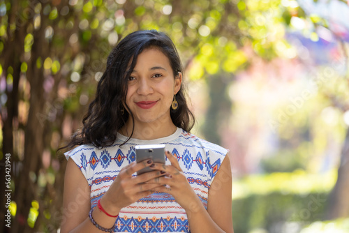 Real Mexican woman holding smart phone and looking at camera outdoors