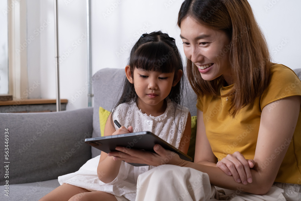 Fototapeta premium Young Asian girl using digital tablet for painting picture or study online lesson at home, with mother support and teaching the lesson inside of living room. Online education or home school concept.