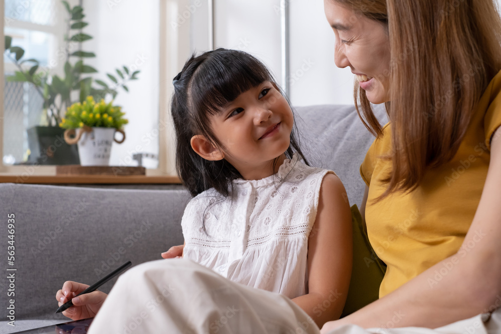 Young Asian girl using digital tablet for painting picture or study online lesson at home, with mother support and teaching the lesson inside of living room. Online education or home school concept. Young Asian girl using digital tablet for painting picture or study online lesson at home, with mother support and teaching the lesson inside of living room. Online education or home school concept.