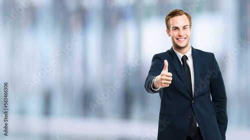 Image of excited businessman showing thumbs up like hand sign gesture  in black suit  over blurred interior background. Handsome happy man. Copy space for ad.