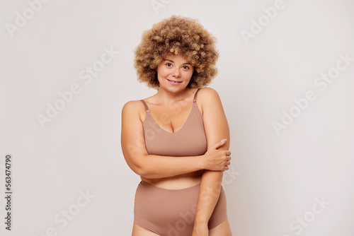 Horizontal shot of overweight adult woman with curly hair hug herself, feels confident and self loved, poses in beige lingerie over white background. Self acceptance and body positivity concept © South House Studio
