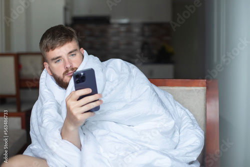 Young lazy sleepy serious man is using his smartphone, cell mobile phone at home in bedroom or living room covered in blanket, looking at screen, reading message or typing