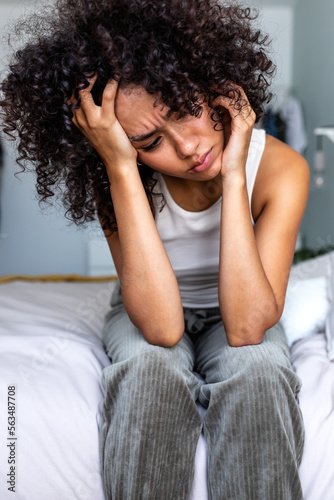 Vertical portrait of young african american latina woman sitting on bed ledge feeling depressed and sad.