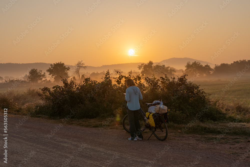 Obraz premium Woman traveling on a bicycle loaded with bags on the side of a dirt road at sunrise in Cambodian countryside