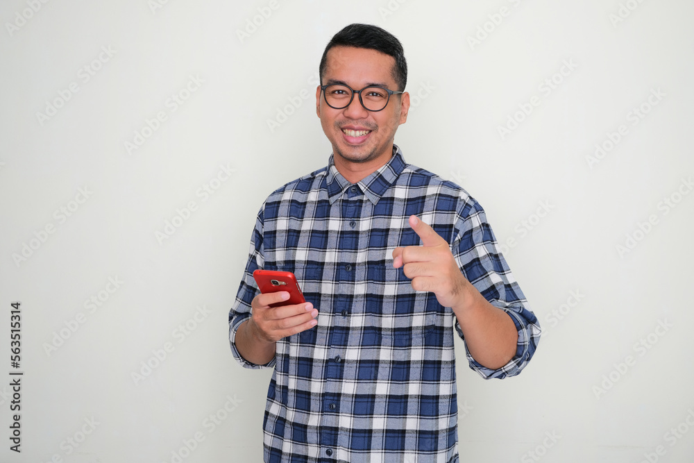Adult Asian man smiling and pointing to the camera while holding mobile phone