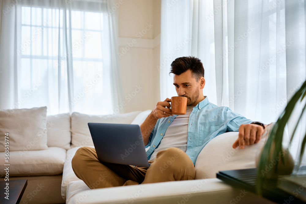 Mid adult man drinks coffee while using laptop and relaxing in the living room. Mid adult man drinks coffee while using laptop and relaxing in the living room.