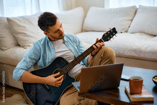 Mid adult man plays acoustic guitar while using laptop at home.