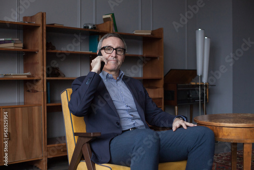 Nervous worried elderly businessman in blue suit and eye glasses talking on his mobile phone on grey wall background