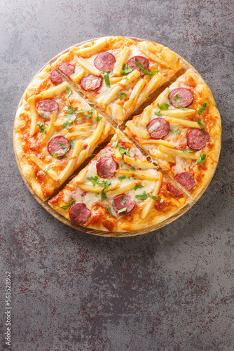 Sliced pizza with french fries, sausages, mozzarella cheese, tomato sauce and herbs close-up on a wooden board on the table. Vertical top view from above