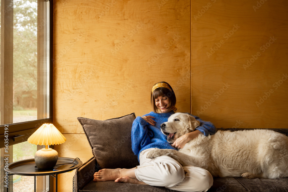 Woman cares her dog while sitting on a couch by the window, spending leisure time together in wooden house on nature