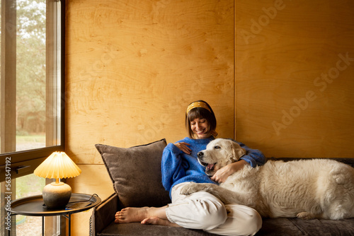 Obraz na płótnie Woman cares her dog while sitting on a couch by the window, spending leisure time together in wooden house on nature w salonie