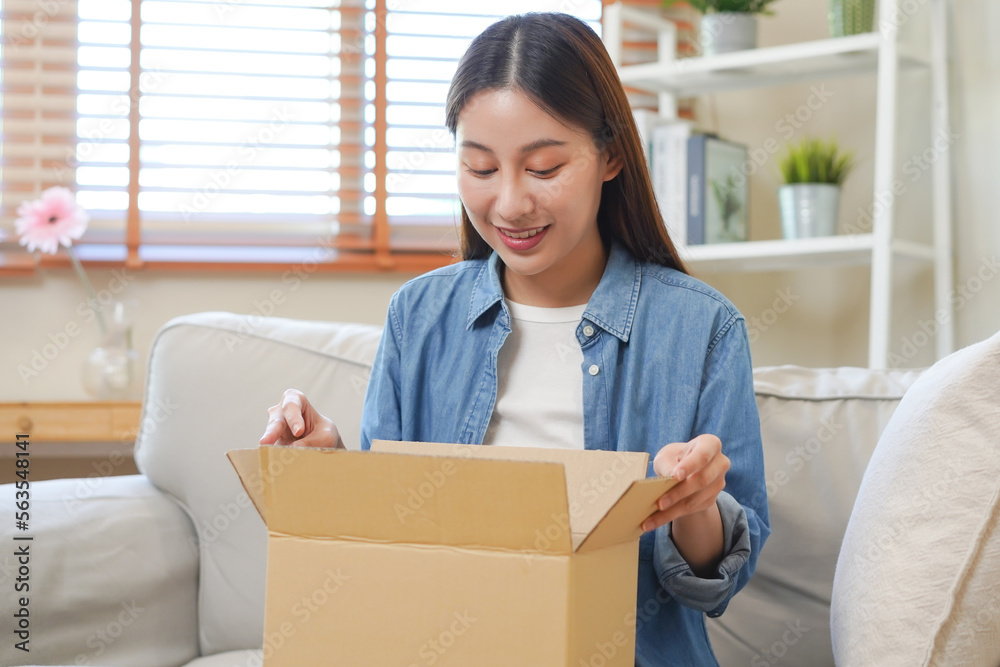 Happy excited, asian young woman, girl customer sitting on sofa at home, opening and unpacking cardboard box carton parcel after buying ordering present, shopping online, delivery service concept. Happy excited, asian young woman, girl customer sitting on sofa at home, opening and unpacking cardboard box carton parcel after buying ordering present, shopping online, delivery service concept.
