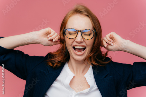 Fototapeta Naklejka Na Ścianę i Meble -  Horizontal shot of ginger woman in formal clothing, demonstrates gesture of ignore, plugs ears, shouts loudly, wears transprent glasses for vision correction, keeps eyes shut, isolated on pink wall