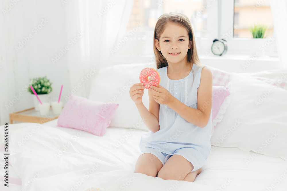 Lovely small girl holds delicious doughnut in hands, going to have breakfast, poses on bed in cozy white bedroom, looks at camera with positive expression. Children, dessert, bed time concept Lovely small girl holds delicious doughnut in hands, going to have breakfast, poses on bed in cozy white bedroom, looks at camera with positive expression. Children, dessert, bed time concept