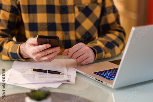 Unrecognizable uses smartphone and laptop. Documents lies on table. Hands close-up. Concept of remote work and freelancing