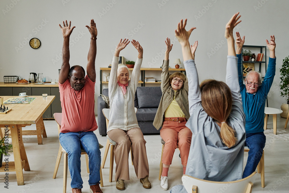 Group of senior people sitting on chairs and repeating exercises instructor during morning