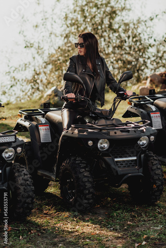 sexy girl with long dark hair on quad bike; dressed in black leather clothes; sunglasses on the eyes; posing on a quad bike