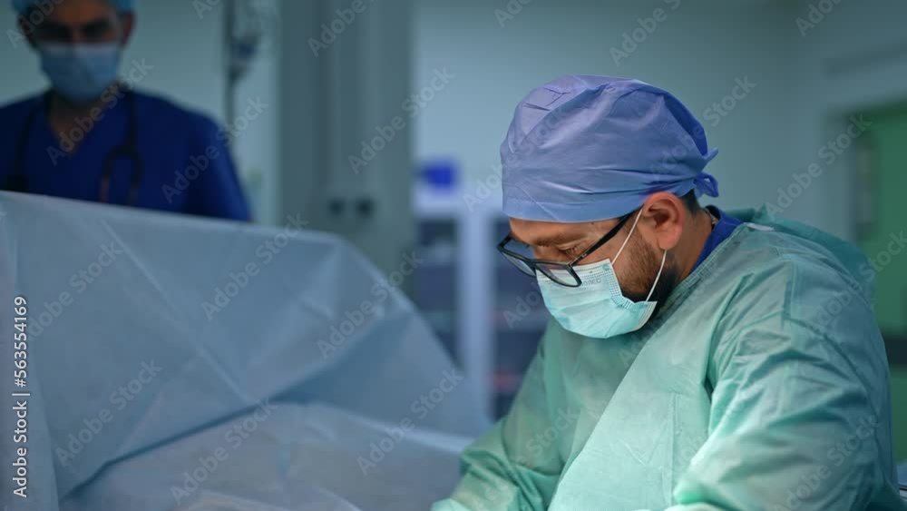 Mid-aged male Caucasian surgeon performing surgery. Anesthesiologist at backdrop in blur.