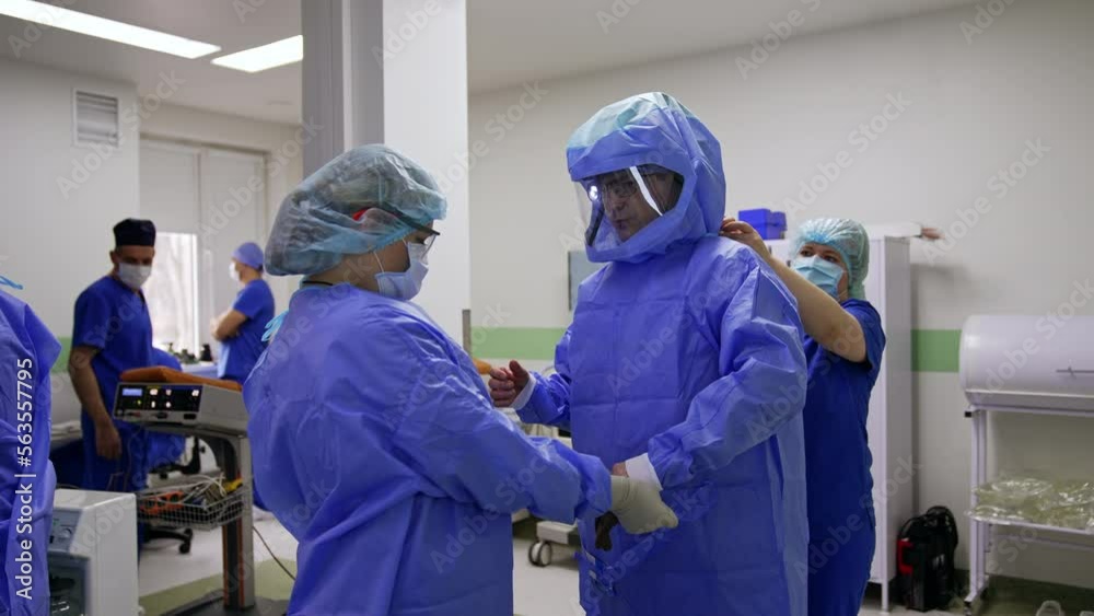 Caucasian doctor in glasses is being dressed in protective suit. Nurses are helping the surgeon to put on robe and gloves. Caucasian doctor in glasses is being dressed in protective suit. Nurses are helping the surgeon to put on robe and gloves.
