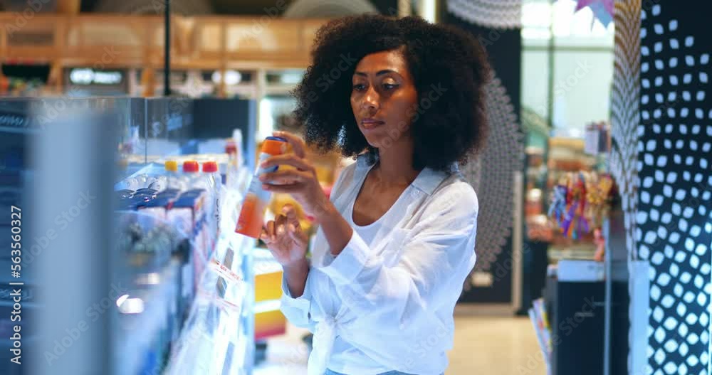 Pleasant African American woman, stands by shef with deodorants assortment, reads the content on label, checks price on the tag while shopping in the hygiene department of supermarket in shopping mall