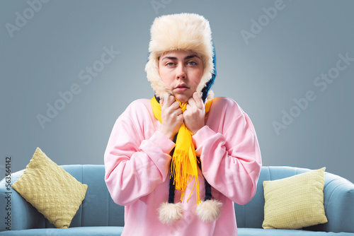 Young freezed woman in ushanka hat and yellow scarf is warming herself. Gray background with sofa. The concept of energy crisis and winter season photo