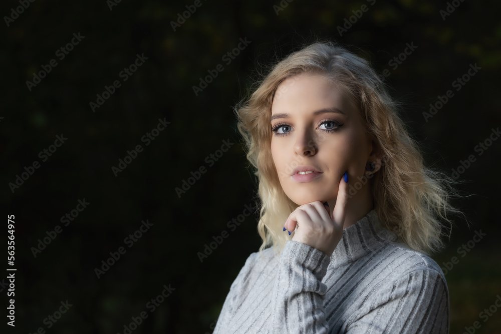 Portrait of attractive young woman dressed in a sweater posing outdoors against a dark background. Horizontally.
