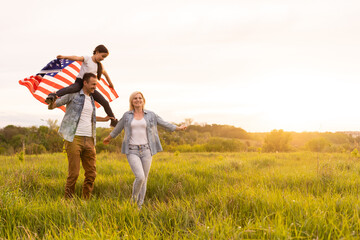 Young parents with their daughter holding American flag in countryside at sunset. Independence Day celebration