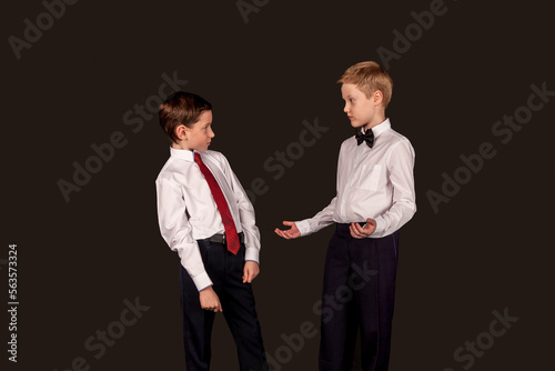 Two confident boys friends in white shirt posing isolated on black background, looking at each other speaking talking. Children debating argument. Business discussion concept. Copy text space for ad