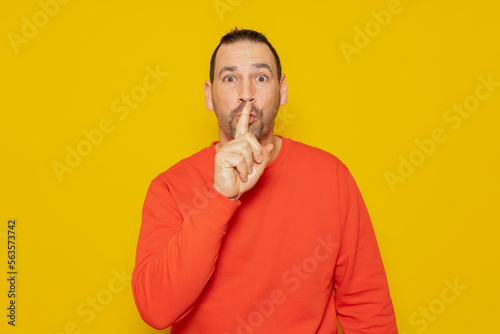 A serious handsome man in a red sweater, puts his index finger to his lips, tries to keep the conspiracy, looks to the side and says: Shh, be quiet please. Isolated shot of man shows silence sign. © Andres