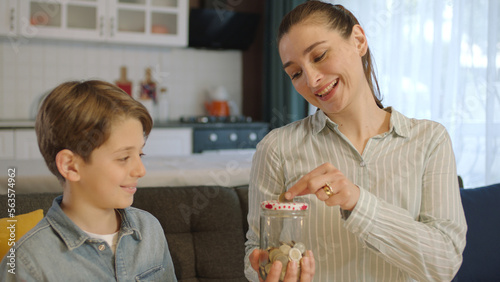 Young mother and her little son putting coins in piggy bank while sitting on sofa at home. The mother teaches the child to be thrifty, to save money, to think about the future, to save. photo