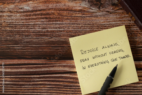 Rejoice always, pray without ceasing, give thanks to God Jesus Christ, handwritten new testament verse (Thessalonians 5:16) on note with pen and holy bible book on wooden table. Copy space, top view.  photo