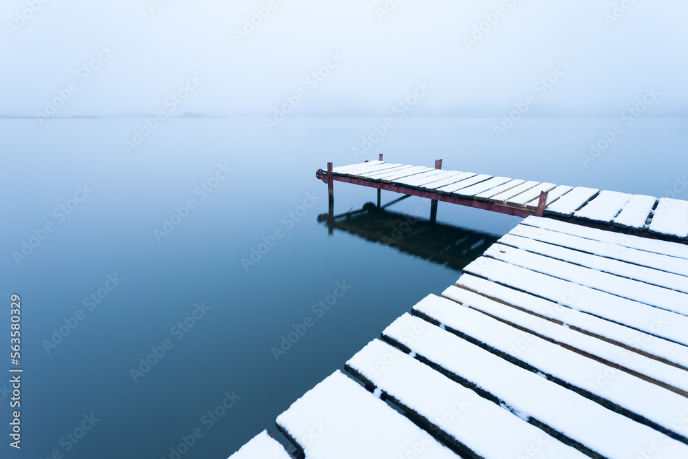 A snow-covered pier with a calm lake on a foggy day