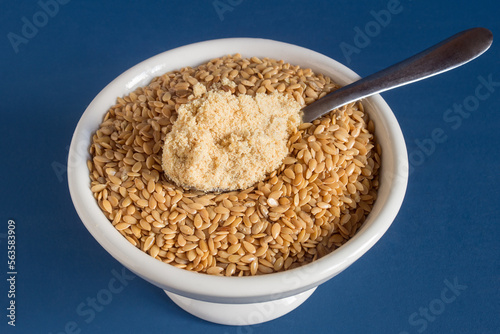 A teaspoon of golden flax powder over a bowl filled with whole flax seeds on a blue studio background. Healthy raw food.
